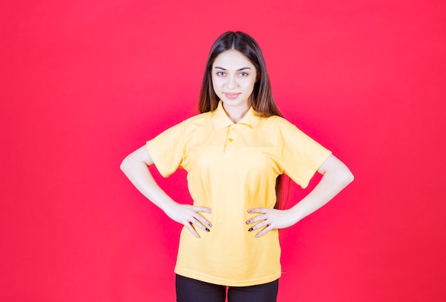 Giovane donna in camicia gialla in piedi sul muro rosso