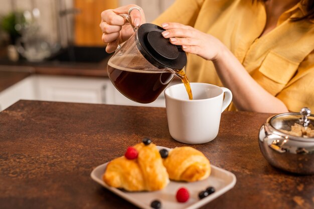 Giovane donna in camicia gialla seduti a tavola e versando una tisana fresca nella tazza mentre sta per avere un croissant fresco per colazione