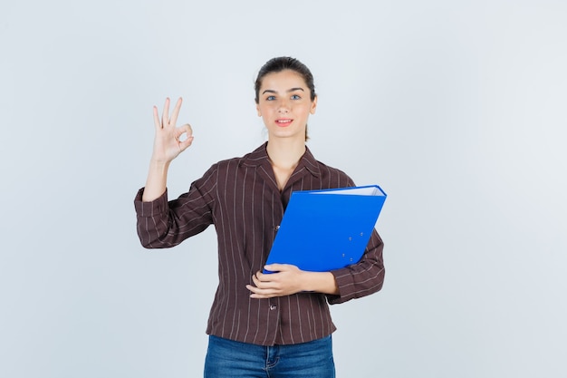 Giovane donna in camicia, jeans con cartella, che mostra gesto ok e sembra premurosa, vista frontale.