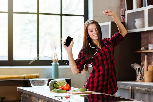 Giovane donna in camicia rossa con gli occhi chiusi ascoltando musica, ballando in cucina con telefono e cuffie.