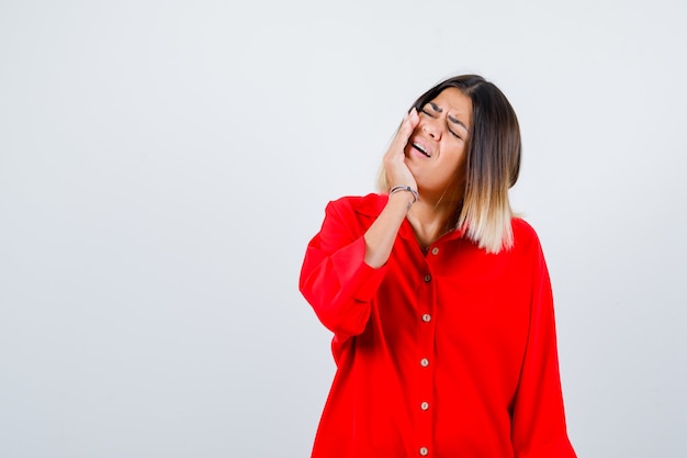 Giovane donna in camicia rossa oversize che soffre di mal di denti e sembra dolorosa, vista frontale.