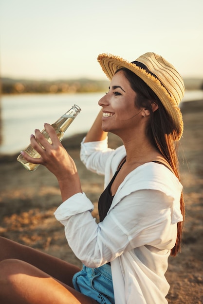 Giovane donna in cappello di paglia che si rilassa al tramonto sulla riva del fiume e beve birra.