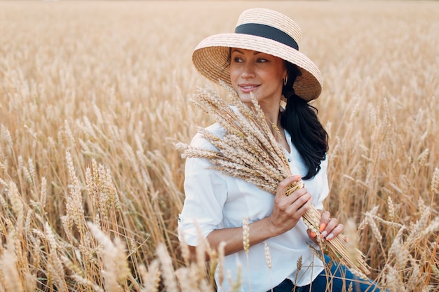 Giovane donna in cappello di paglia che tiene un covone di spighe di grano in campo agricolo.