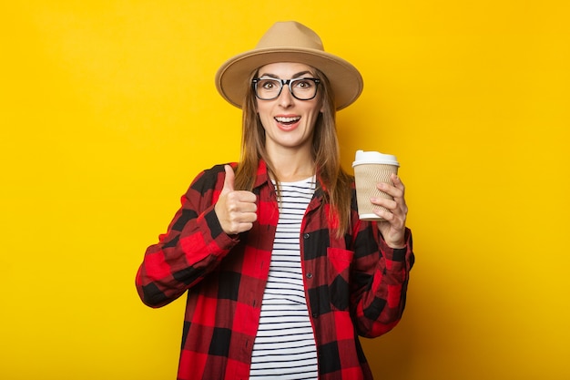 Giovane donna in cappello e camicia a quadri che tiene tazza di carta con caffè e che fa gesto di mano su colore giallo