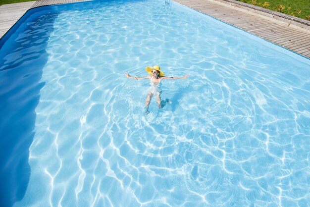 Giovane donna in cappello giallo che nuota nel bacino con acqua blu. Vista generale dall'alto