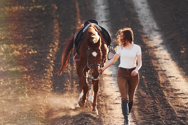 Giovane donna in cappello protettivo che cammina con il suo cavallo nel campo agricolo durante la giornata di sole