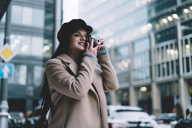 giovane donna in cappotto e cappello in piedi e scattare foto sulla fotocamera mentre sorride sullo sfondo