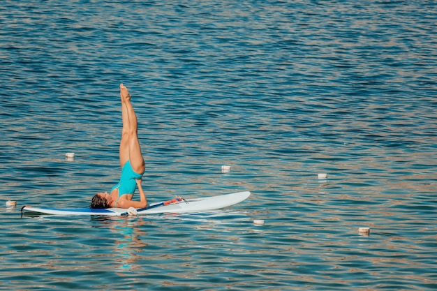 Giovane donna in costume da bagno blu che fa yoga su una tavola con la pagaia