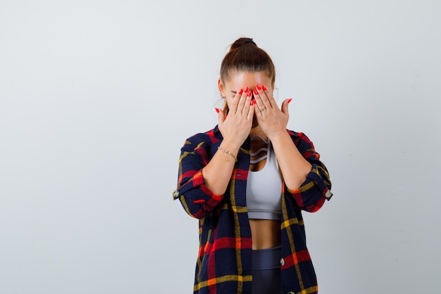 Giovane donna in crop top, camicia a scacchi, pantaloni con le mani sul viso e sguardo malinconico, vista frontale.