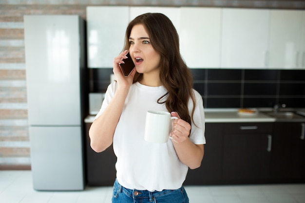 Giovane donna in cucina. Emozionalmente parlando al telefono e meraviglia. Tenere la tazza bianca con drink in mano.