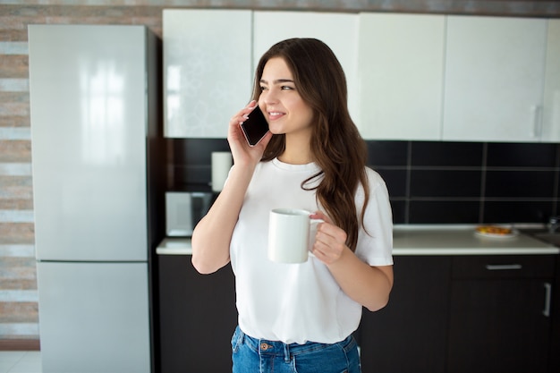 Giovane donna in cucina. Parlare su smartphone e sorridere. Periodo delle moderne tecnologie. Lavorare online.