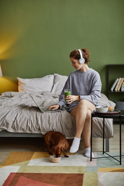 Giovane donna in cuffia che comunica online sul laptop durante la colazione in camera da letto