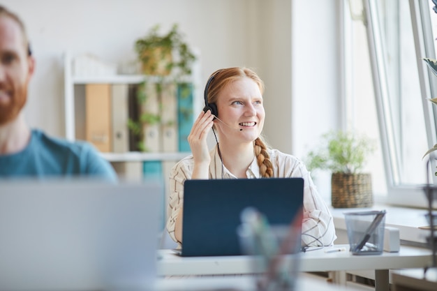 Giovane donna in cuffie seduto al tavolo con il computer portatile che lavora nel call center