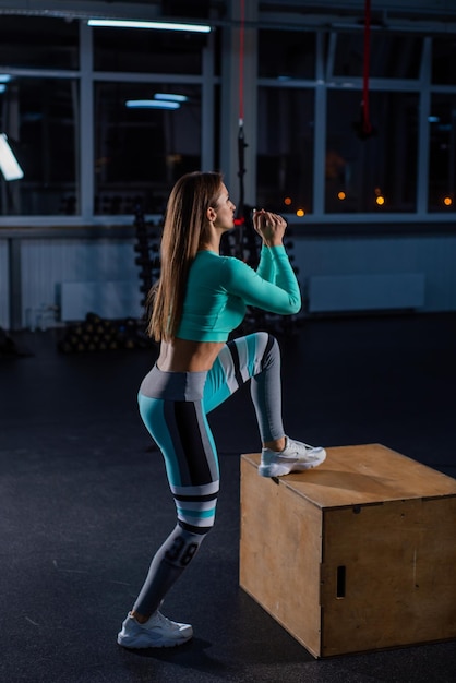 Giovane donna in forma che fa box jumping su sfondo scuro con spazio di copia Salto alto su un piede su una scatola Bella atleta esegue un allenamento circolare Accovacciarsi