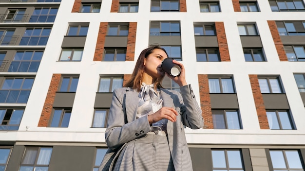 Giovane donna in giacca e cravatta beve caffè da un bicchiere di plastica sullo sfondo di un edificio in una giornata di sole