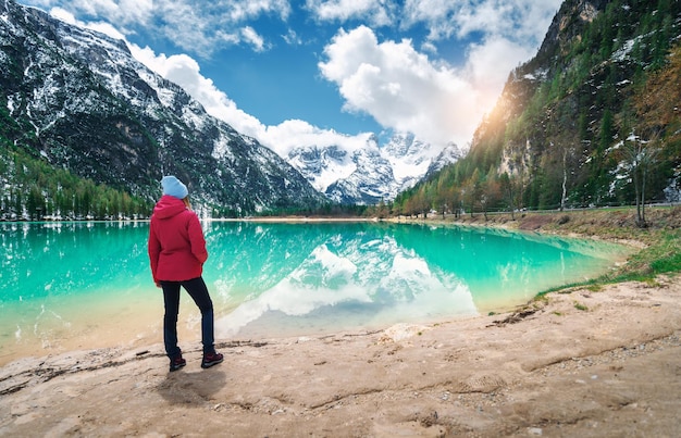 Giovane donna in giacca rossa è in piedi sulla riva del lago con acqua azzurra in una giornata di sole in primavera Paesaggio con il riflesso della ragazza nell'acqua montagne innevate cielo blu con nuvole alberi verdi Viaggio