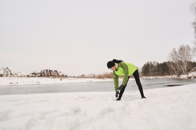 Giovane donna in giacca verde in piedi t lago invernale e chinarsi mentre fa esercizio di riscaldamento