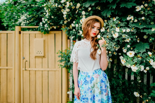 Giovane donna in gonna lunga vintage, top vecchio stile con capelli rossi ricci e cappello di paglia in piedi tra i cespugli con recinzione in legno