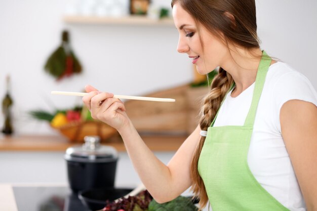 Giovane donna in grembiule verde sta cucinando in una cucina La casalinga sta assaggiando la zuppa con un cucchiaio di legno