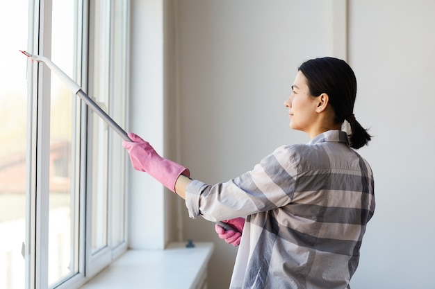 Giovane donna in guanti che puliscono la finestra con la scopa in ufficio