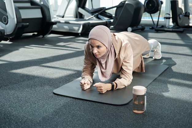 Giovane donna in hijab e abbigliamento sportivo che fa uno sforzo mentre fa la tavola sul tappetino nel centro fitness o in palestra