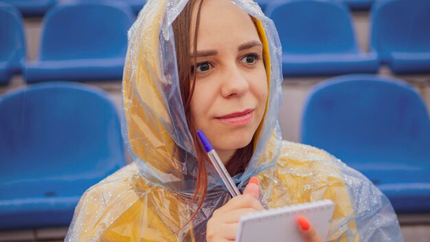 Giovane donna in impermeabile con penna blocco note seduta sulle gradinate dello stadio da sola in caso di pioggia Giornalista che scrive note durante l'allenamento sportivo allo stadio di strada