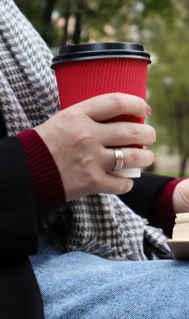 Giovane donna in jeans, cappotto e sciarpa, su una panchina del parco. Una donna sta leggendo un libro e bevendo caffè o altra bevanda calda all'aperto da sola. Avvicinamento. Il concetto di onore, studio, tempo libero e ricreazione.