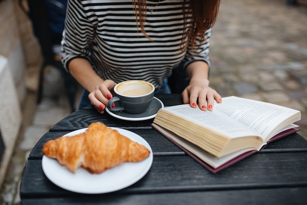 Giovane donna in maglione a righe che legge un nuovo libro mentre riposa sulla terrazza del caffè