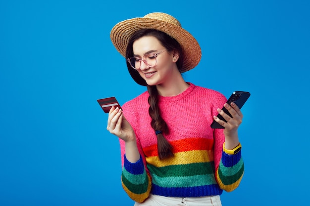 Giovane donna in maglione arcobaleno in possesso di carta di credito in plastica e telefono cellulare