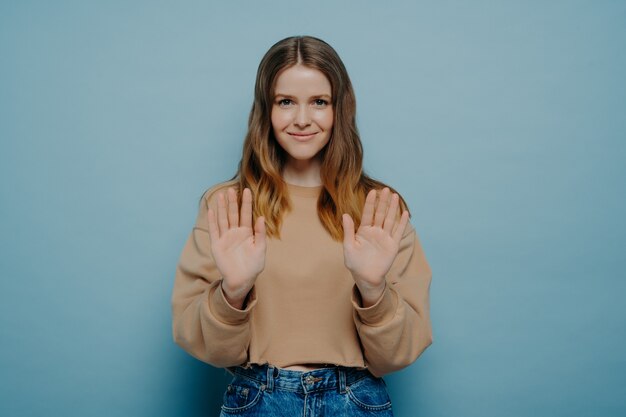 Giovane donna in maglione beige che esprime disaccordo, mostrandolo tenendosi per mano di fronte a lei, dicendo di no con il suo gesto, rifiutandosi educatamente di fare ciò che ha chiesto su sfondo azzurro