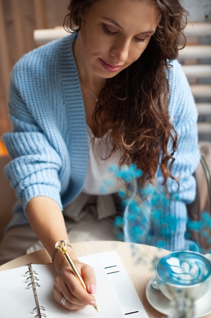 Giovane donna in maglione caldo blu che si siede vicino alla grande finestra della caffetteria e scrive una lista della spesa di Natale con una tazza di latte blu. Pianificare le vacanze di Natale. Concetto di organizzazione e pianificazione.