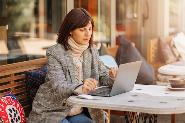 Giovane donna in maglione caldo e blazer che lavora al net-book nella caffetteria sulla terrazza. Mattina d'autunno. Stile di strada.