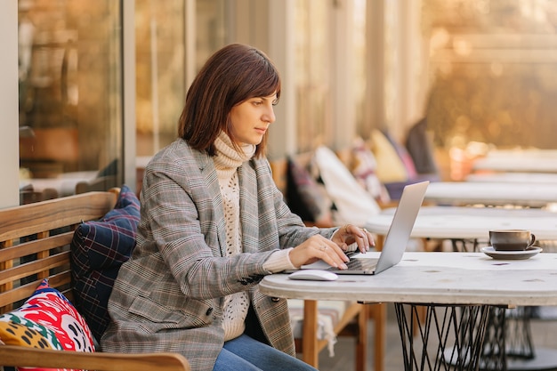Giovane donna in maglione caldo e blazer che lavora al net-book nella caffetteria sulla terrazza. Mattina d'autunno. Stile di strada.