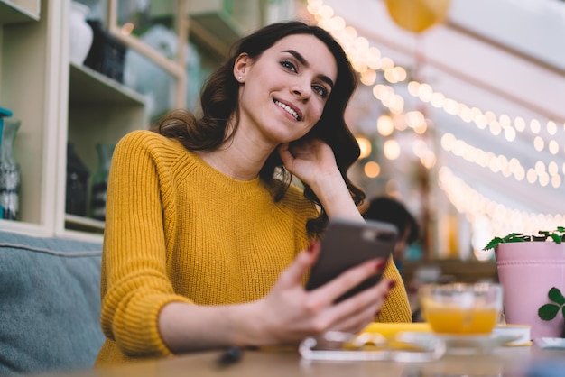 giovane donna in maglione giallo con capelli castani e smartphone che si tocca i capelli e si siede alla scrivania