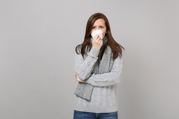 Giovane donna in maglione grigio, sciarpa con naso che cola, soffiando il naso al tovagliolo isolato su sfondo grigio muro. Stile di vita sano, trattamento delle malattie malate, concetto di stagione fredda. Mock up copia spazio.