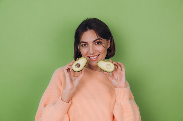 Giovane donna in maglione pesca casual isolato sulla parete verde oliva che tiene avocado, salute e concetto di cura della pelle, copia dello spazio