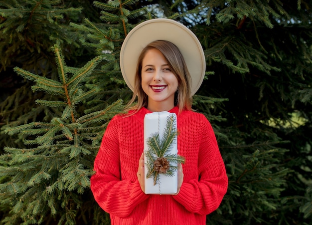 Giovane donna in maglione rosso con regalo di Natale vicino all'albero di pino
