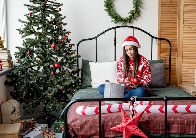 Giovane donna in maglione rosso e cappello da Babbo Natale seduto sul letto che celebra il natale