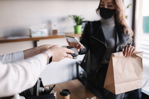 Giovane donna in maschera di pagare la bolletta tramite smartphone utilizzando la tecnologia nfc in un cliente femminile di caffè