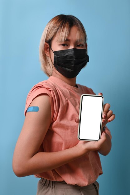 Giovane donna in maschera facciale che mostra il braccio vaccinato e tiene in mano uno smartphone