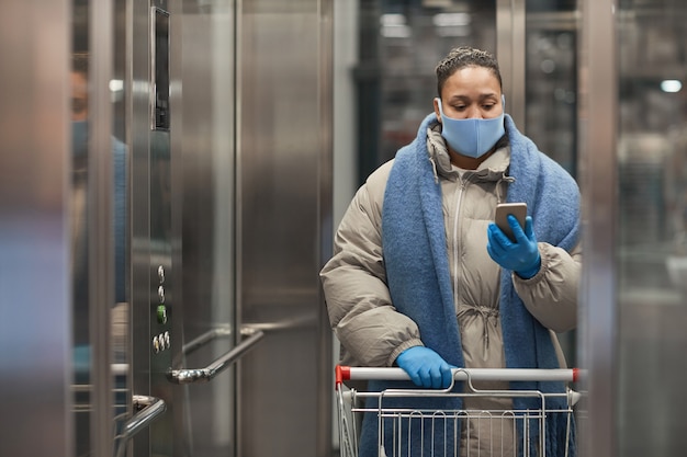 Giovane donna in maschera protettiva e guanti che fanno selfie ritratto sul suo telefono con il carrello in ascensore