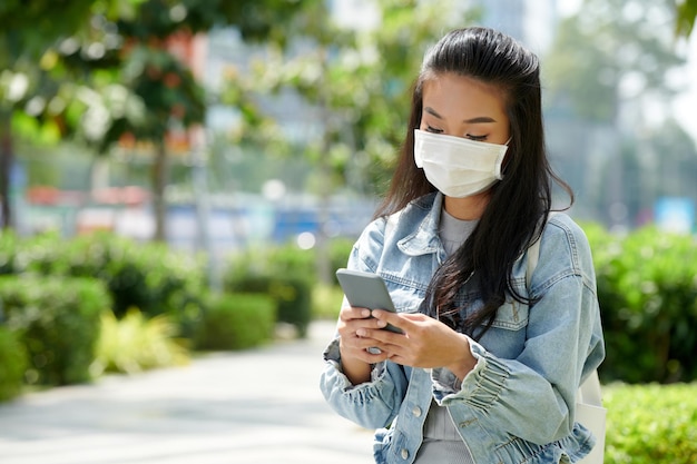 Giovane donna in maschera protettiva seduta nel parco e sms agli amici