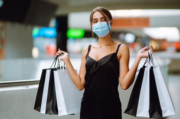 Giovane donna in mascherina medica sterile protettiva sul viso con borse della spesa nel centro commerciale.