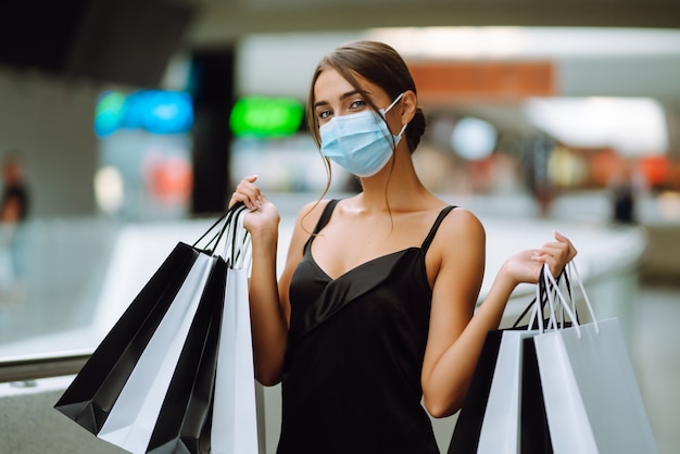 Giovane donna in mascherina medica sterile protettiva sul viso con borse della spesa nel centro commerciale.