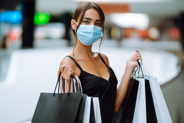 Giovane donna in mascherina medica sterile protettiva sul viso con borse della spesa nel centro commerciale.