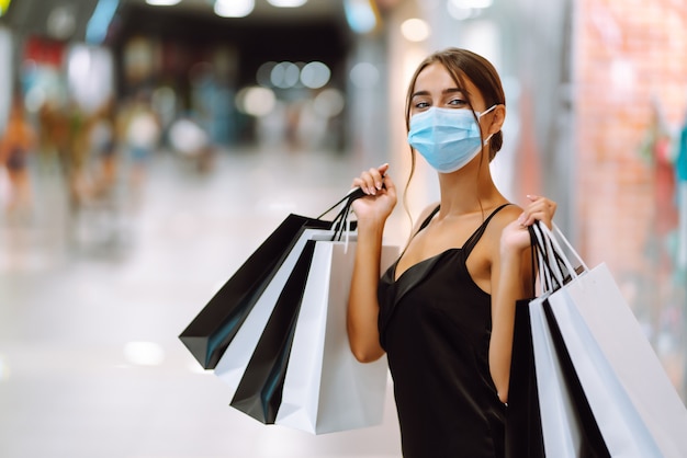 Giovane donna in mascherina medica sterile protettiva sul viso con borse della spesa nel centro commerciale.