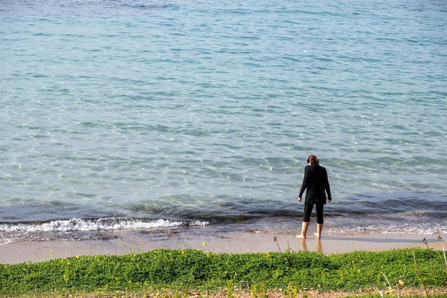 Giovane donna in nero a piedi nudi si trova sulla spiaggia sabbiosa greca in attesa che l'onda del mare le bagni la gamba