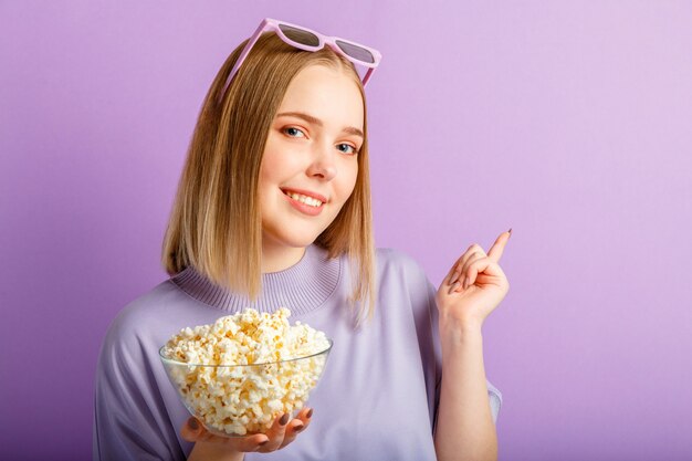 Giovane donna in occhiali da cinema che guarda film in 3d. Spettatore di film sorridente ragazza adolescente in bicchieri con spettacolo di popcorn con la mano sullo spazio della copia isolato su sfondo viola