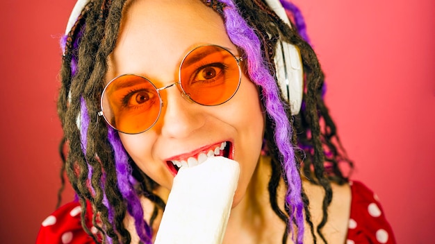 Giovane donna in occhiali da sole cuffie mordere il gelato sorridente guardando la fotocamera Femmina allegra con dreadlocks mangiare gelato ascoltando musica in studio