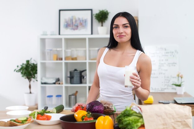 Giovane donna in piedi accanto al tavolo da cucina con verdure fresche che bevono latte al mattino